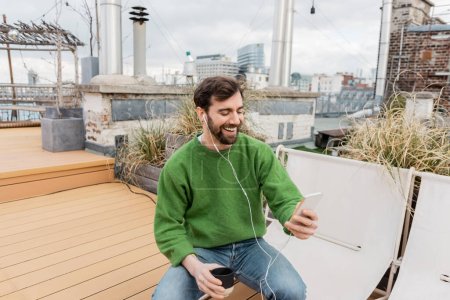 Cheerful man in earphones holding cup of coffee while spending time on rooftop terrace in Vienna