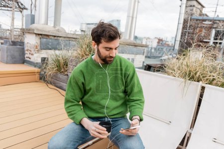 bearded man listening music in earphones and holding cup of coffee on rooftop in Vienna, Austria