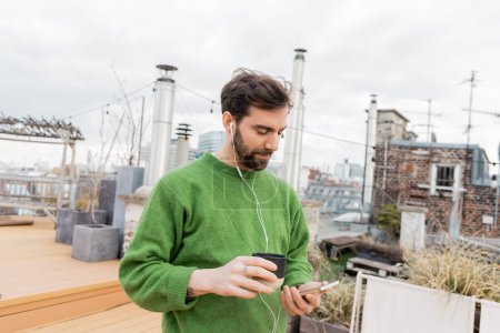 handsome man listening music in earphones and holding cup of coffee on rooftop in Vienna, Austria