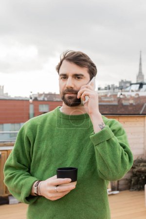 Mann im Pullover spricht mit Smartphone und hält Tasse Kaffee auf Dach in Wien