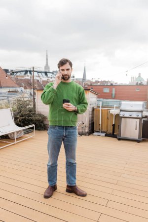 bärtiger Mann im Pullover, der auf dem Dach in Wien mit dem Smartphone spricht und Kaffee trinkt