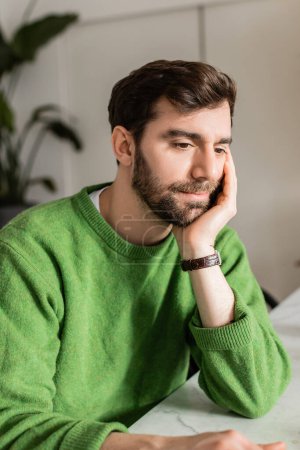 Retrato de hombre barbudo en jersey verde y reloj de pulsera mirando hacia otro lado mientras descansa en casa  