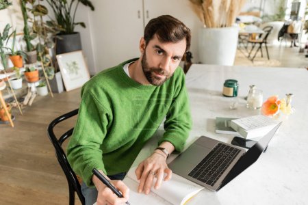 Foto de Freelancer barbudo en jersey verde mirando la cámara mientras escribe en el cuaderno en casa - Imagen libre de derechos
