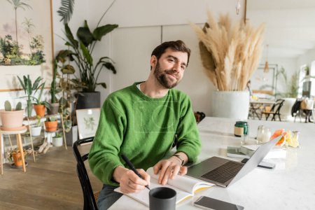 Téléchargez les photos : Freelance souriant et barbu en pullover vert écrit sur ordinateur portable près des appareils sur la table - en image libre de droit
