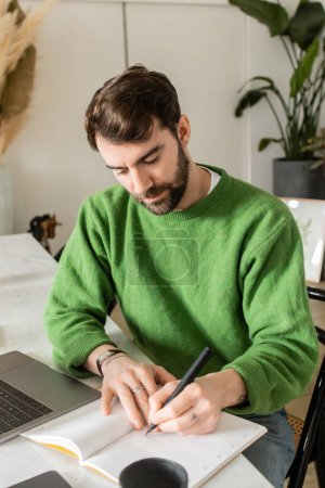 Foto de Freelancer barbudo en jersey verde escrito en portátil cerca de laptop, smartphone y taza de café - Imagen libre de derechos
