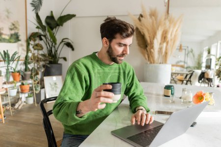 Téléchargez les photos : Entrepreneur barbu en pull vert tenant une tasse de café et utilisant un ordinateur portable à la maison - en image libre de droit