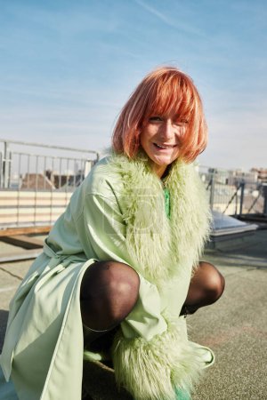 mujer alegre y de moda sentada en las horcas y mirando a la cámara en la terraza de la azotea en Viena