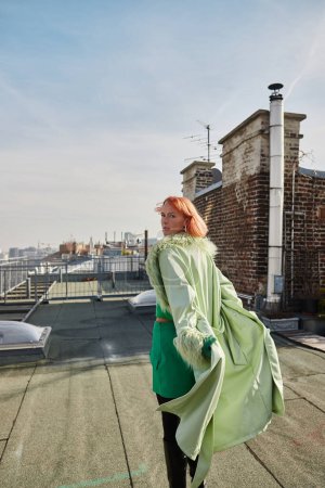 attractive trendy woman looking at camera and walking on roof terrace in Vienna, street fashion