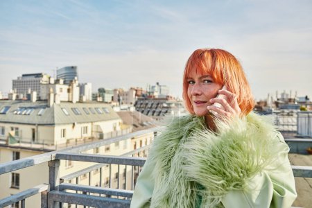 mujer de moda positiva hablando en el teléfono inteligente y mirando a la cámara en la terraza de la azotea en Viena, Austria