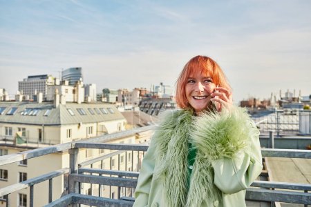 Eine lächelnde, stylische Frau, die auf dem Smartphone spricht und auf der Dachterrasse in Wien wegschaut