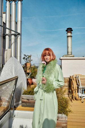 Trendfrau in stylischer Kleidung hält Blumen in die Kamera auf der Dachterrasse in Wien
