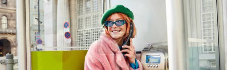Foto de Mujer con estilo en gafas de sol y chaqueta de piel sintética sonriendo en cabina telefónica en la calle en Viena, pancarta - Imagen libre de derechos