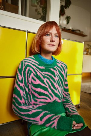 dreamy woman in trendy animal print sweater sitting and looking away in modern apartment in Vienna