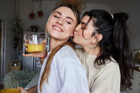Téléchargez les photos : Couple lesbien affectueux dans les années 20 partageant un baiser, femme tenant du jus frais dans une cuisine confortable - en image libre de droit
