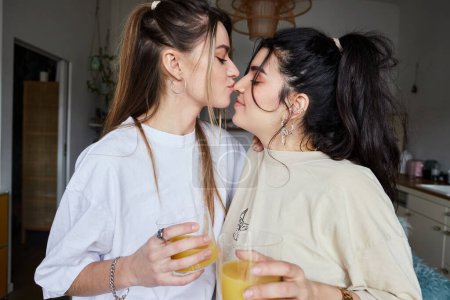 Téléchargez les photos : Jeune femme tenant un verre de jus d'orange et embrassant nez de petite amie dans la cuisine, couple lgbt - en image libre de droit