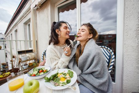 glückliche junge lesbische Frau füttert ihre Freundin beim Frühstück auf dem Balkon, lgbt paar