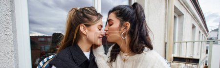 young lesbian woman kissing nose of her girlfriend while sitting together on balcony, lgbt banner