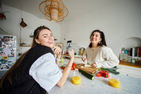 happy young lesbian woman in 20s taking photo of her girlfriend while cooking salad in kitchen