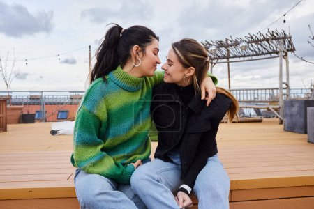 Foto de Momento tierno entre mujeres lesbianas alegres sentadas juntas en una azotea, telón de fondo de paisaje urbano - Imagen libre de derechos