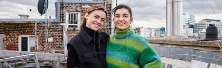 happy lesbian couple in casual attire standing together of rooftop and looking at camera, banner