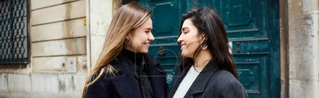 pancarta, feliz pareja lesbiana en abrigos de pie juntos y mirándose el uno al otro en la calle urbana