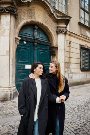 fröhliches lesbisches Paar in Mänteln, das auf der Straße in einer europäischen Stadt zusammensteht und Händchen hält