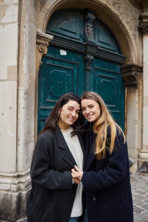 cheerful lesbian couple in coats standing together and holding hands outdoors, looking at camera