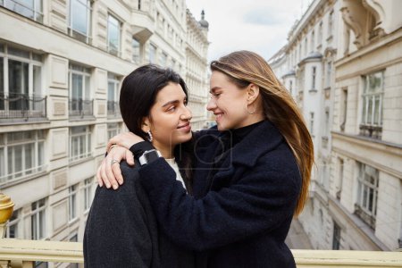 Intimate moment on balcony with city architecture view as backdrop, cheerful lesbian women in love