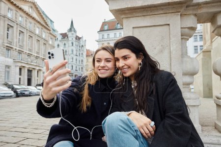 happy and young lesbian couple taking selfie on smartphone while sitting together outdoors in Vienna