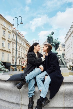 Loving lesbian couple holding hands and sitting by a fountain statue with cityscape in Vienna