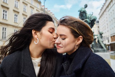 cheerful lesbian couple kissing and sitting by a fountain statue with cityscape in Vienna