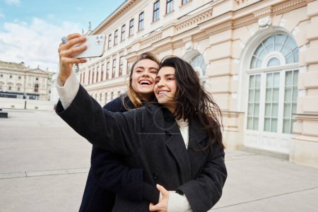 feliz pareja de lesbianas tomando selfie en el teléfono inteligente mientras están de pie juntos cerca del edificio en Viena