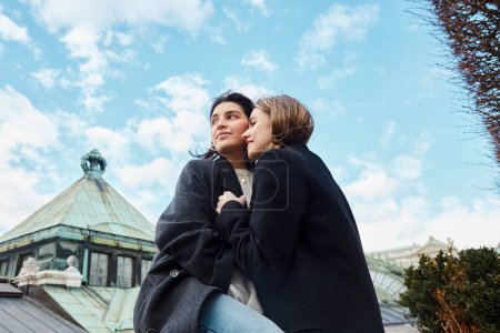 cheerful lesbian couple in outerwear smiling while hugging each other near building in Vienna