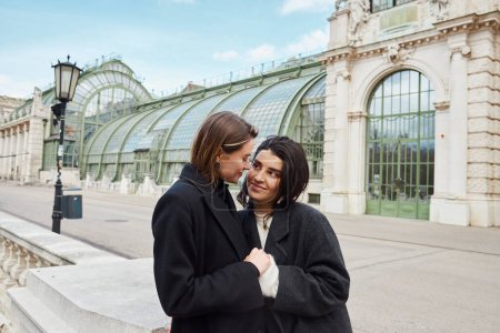 cheerful lesbian couple in coats holding hands of each other near Palmenhaus in Vienna on background