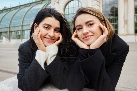 Fröhliches lesbisches Paar in Mänteln blickt im Hintergrund in die Kamera beim Palmenhaus in Wien