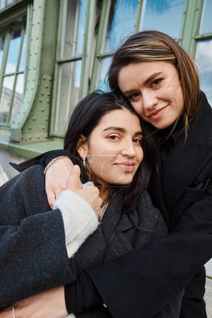portrait of cheerful lesbian couple in coats embracing each other near Palmenhaus in Vienna