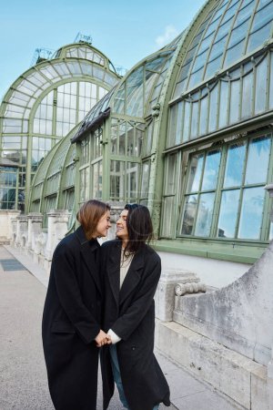 pleased lesbian couple in coats holding hands while standing near Palmenhaus in Vienna on background