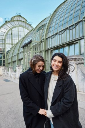 Téléchargez les photos : Couple lgbt en manteaux noirs tendance tenant la main tout en riant près de Palmenhaus à Vienne - en image libre de droit