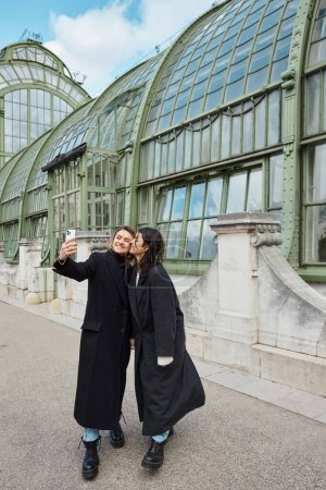 feliz pareja de lesbianas tomando selfie en el teléfono inteligente, mientras que de pie cerca de lugar histórico en Viena