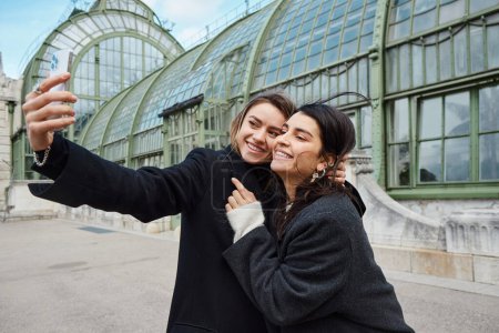 feliz pareja de lesbianas tomando selfie en el teléfono inteligente, mientras que de pie cerca de lugar histórico en Viena