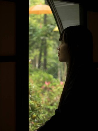 Foto de Side portrait of thoughtful attractive woman looking at green background outside through hotel room or house window - Imagen libre de derechos