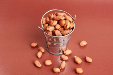 A small metal bucket filled with raw peanuts on a brown background.