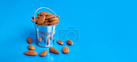 A small metal bucket filled with dried almond kernels on a blue background.