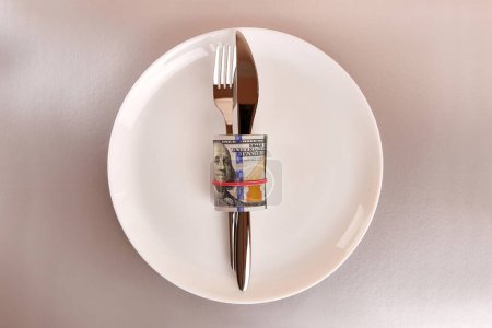 A white plate with a roll of dollars, a fork and a knife on a gray background.
