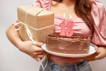 Das Mädchen hält eine festliche Torte mit einer Kerze in Form der Zahl 18 und einem Geschenk in der Hand. Konzept zur Geburtstagsfeier.