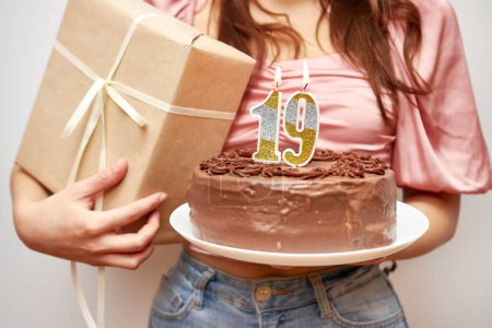 Das Mädchen hält eine festliche Torte mit einer Kerze in Form der Zahl 19 und einem Geschenk in der Hand. Konzept zur Geburtstagsfeier.
