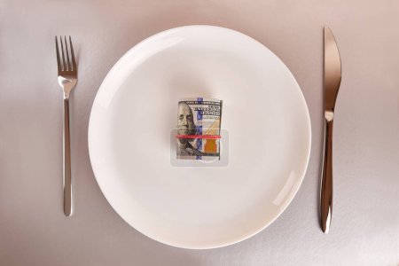 Foto de A white plate with a roll of dollars, a fork and a knife on a gray background. - Imagen libre de derechos