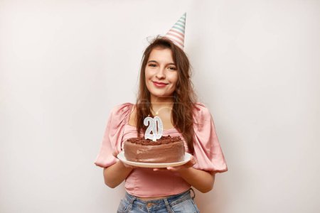 Das Mädchen hält eine festliche Torte mit einer Kerze in Form der Zahl 20. Das Konzept einer Geburtstagsfeier.