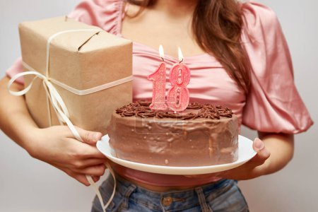 Das Mädchen hält eine festliche Torte mit einer Kerze in Form der Zahl 18 und einem Geschenk in der Hand. Konzept zur Geburtstagsfeier.