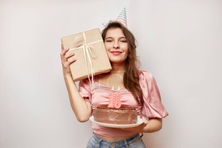 Foto de The girl is holding a festive cake with a candle in the form of the number 18 and a gift. Birthday celebration concept. - Imagen libre de derechos